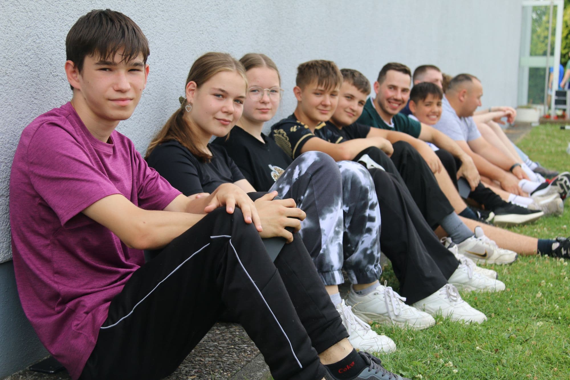 Jugendliche sitzen nebeneinander an einer Hauswand im Gras und lächeln in die Kamera