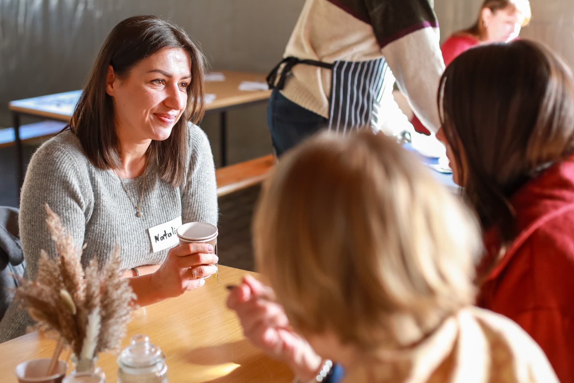 Eine Frau mit Kaffeebecher unterhält sich lächelnd mit anderen am Tisch